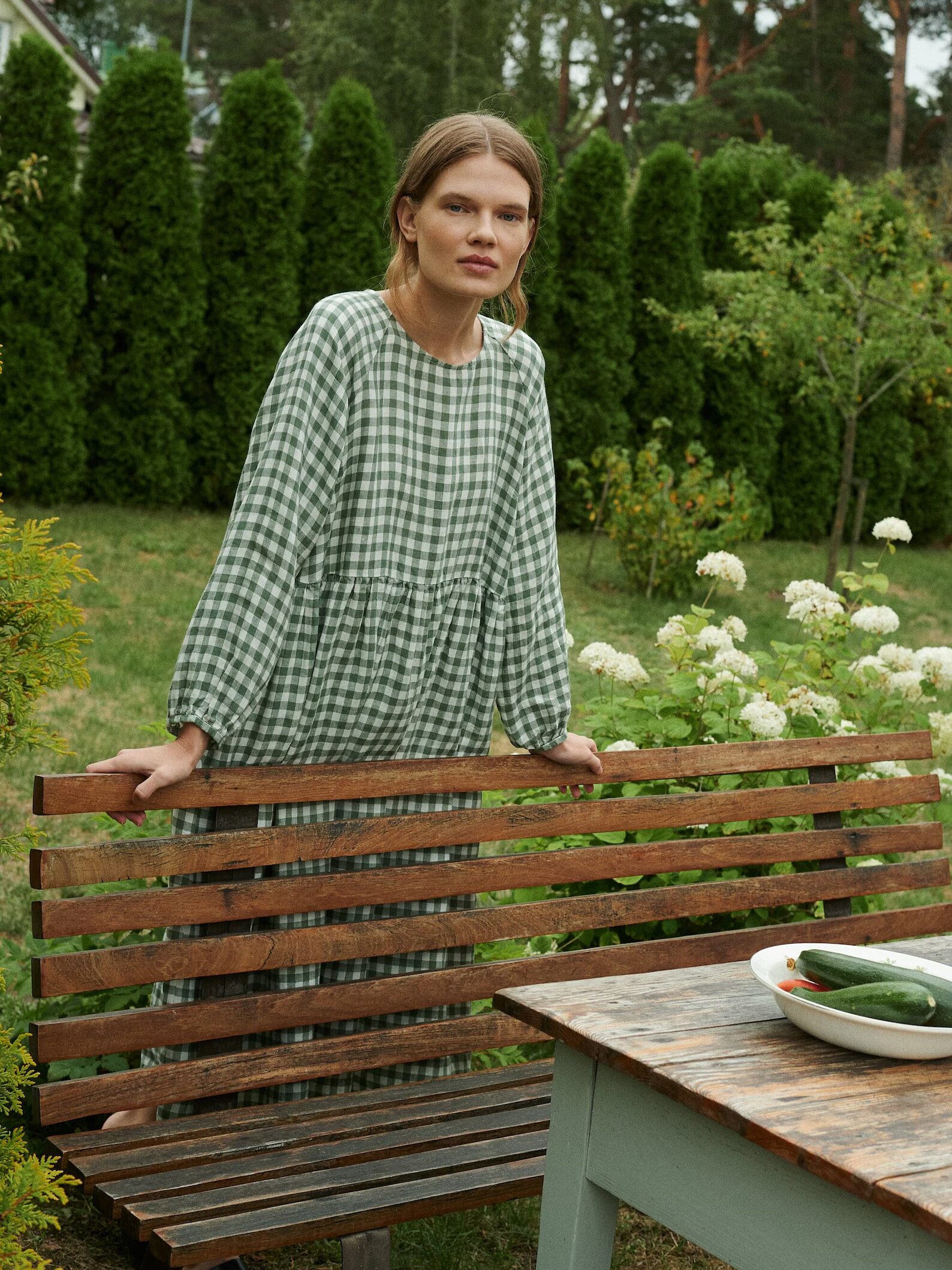 A model leans behind a wooden bench, wearing a green gingham print dress from Linenfox. 