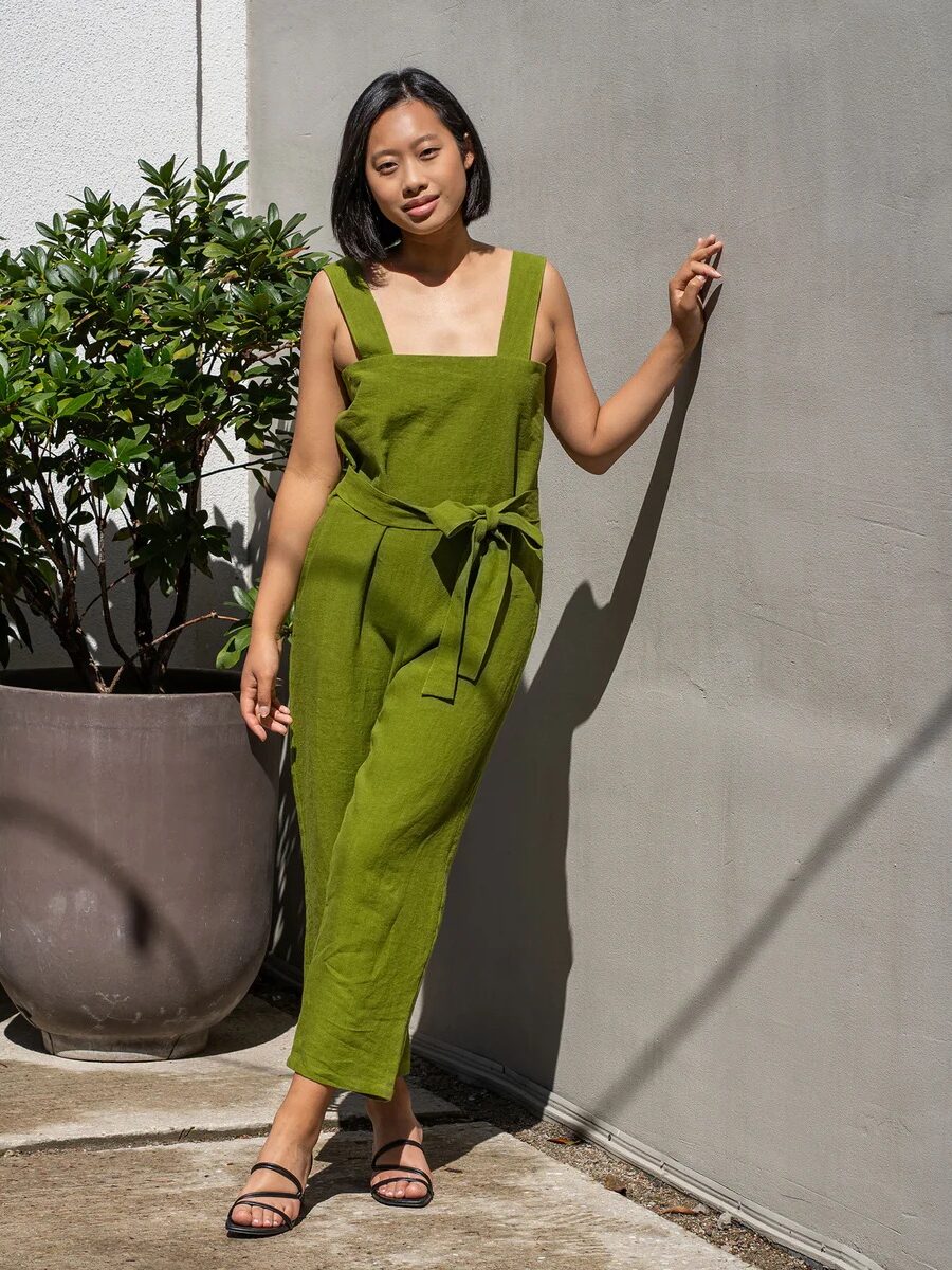 A woman in a green jumpsuit stands by potted plants, touching a wall, sunlight casting shadows around her.