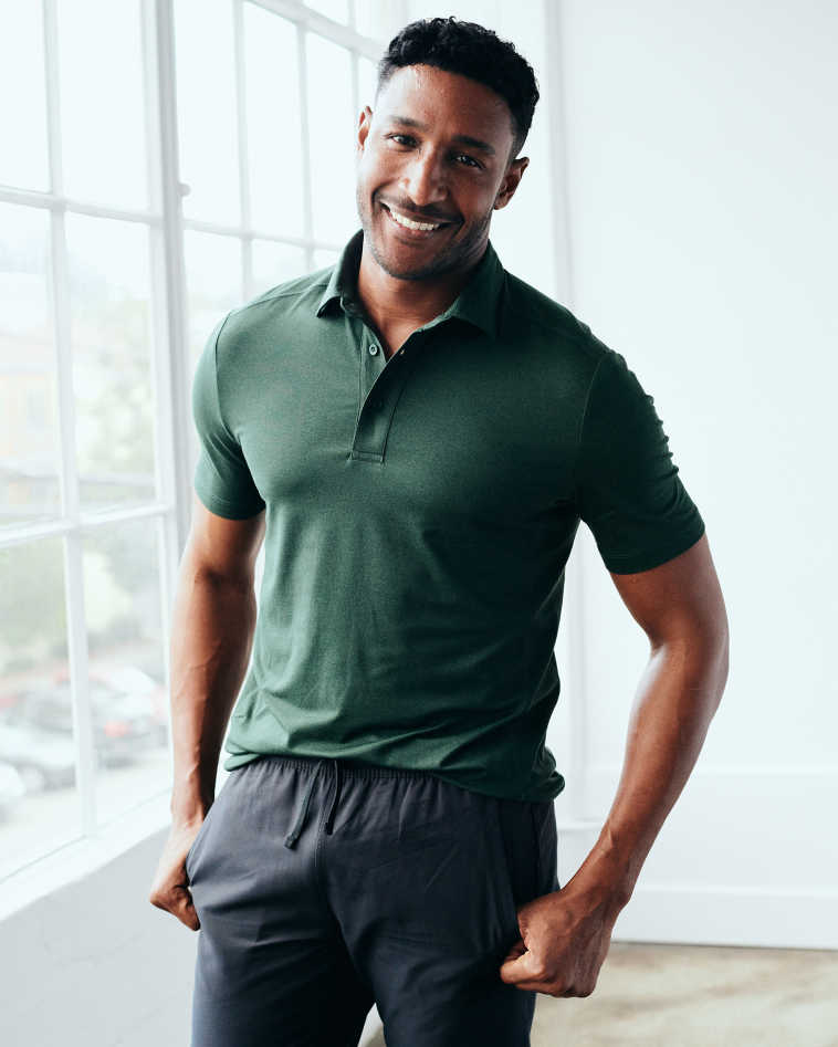 A man in a green polo shirt and black pants stands smiling by a window with natural light.