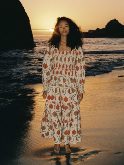 A woman in a floral dress stands on a beach at sunset, with the sun low in the sky between rocks in the background.