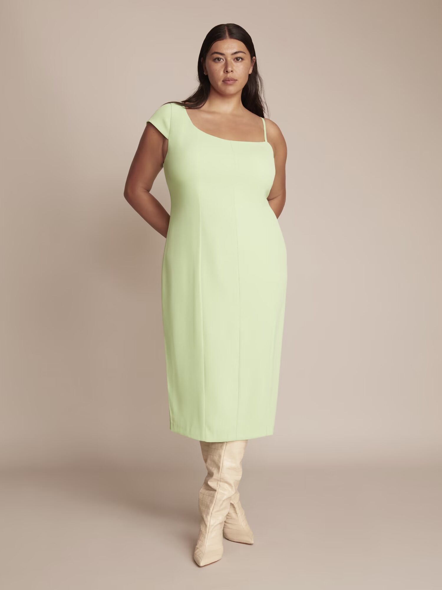 A woman with long dark hair wears a light green dress and beige knee-high boots, posing against a neutral background.