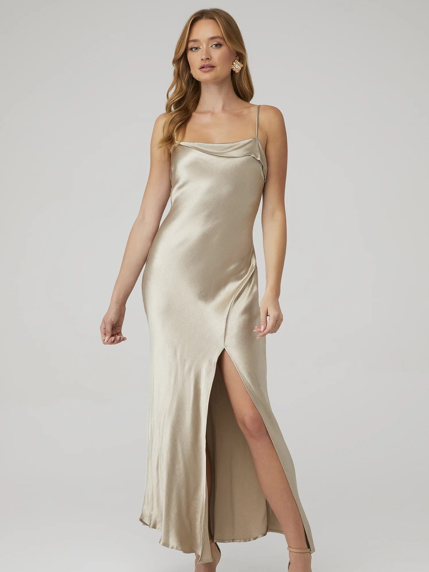 Woman in a silky champagne-colored slip dress with a thigh-high slit, standing against a grey background.