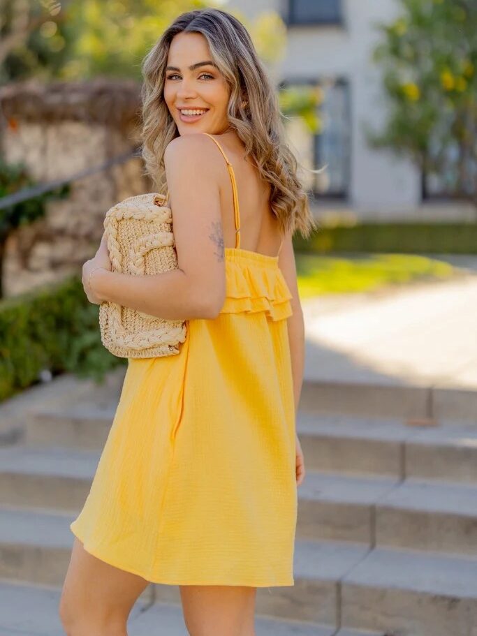 Woman in yellow dress smiling over her shoulder, holding a woven bag, walking outdoors in a sunny, residential area.
