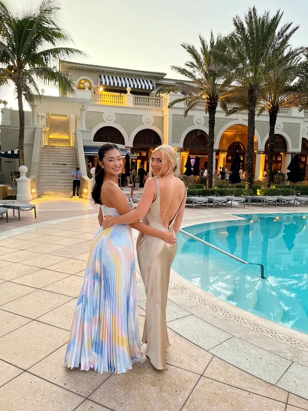 Two women in elegant dresses smiling and posing together at an outdoor event near a pool with palm trees and a building in the background.