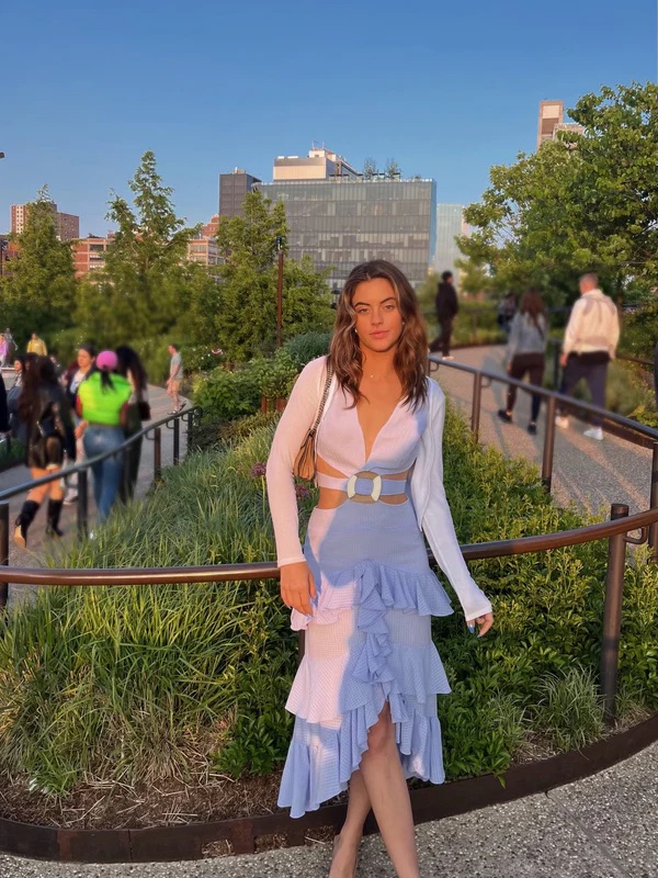 A woman in a stylish blue and white dress and white cardigan stands on a city park path at dusk, with people and buildings in the background.
