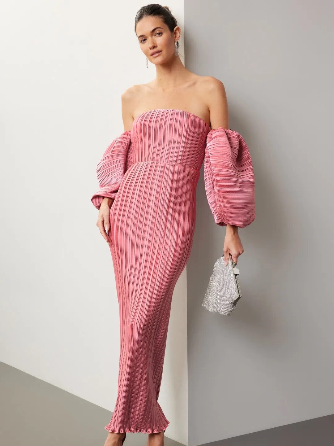 A woman in an elegant pink striped off-the-shoulder gown with puff sleeves, holding a silver clutch, against a light gray backdrop.