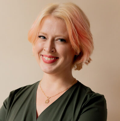 A smiling person with short, light-colored hair and a dark top poses against a plain background.