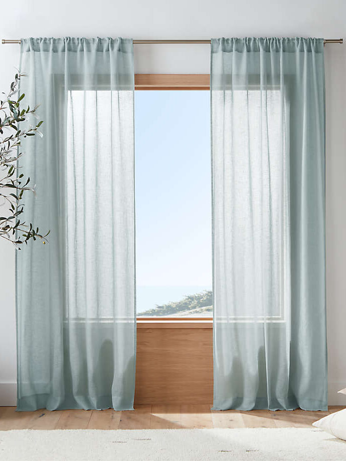 Light blue sheer curtains hang from a rod on a window overlooking a scenic view, with a plant and cushions nearby, in a bright, serene room.