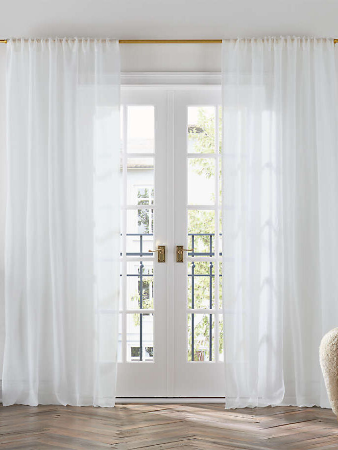 Sheer white curtains hanging on a gold curtain rod, framing a glass door that opens to a balcony, in a room with wooden flooring and a cream armchair.