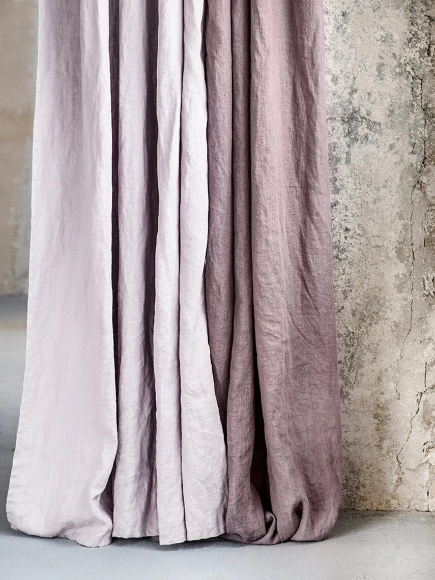 Long, purple curtains drape against a rough-textured wall next to a simple copper wall clock in a minimalist setting.