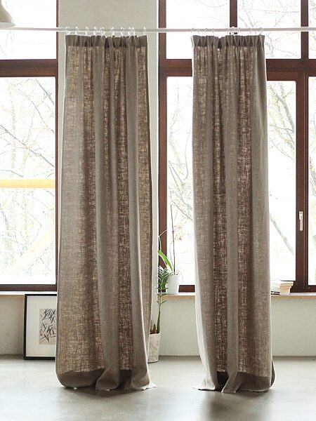Two long, beige curtains hang from a rod in a room with large windows and minimalistic decor.