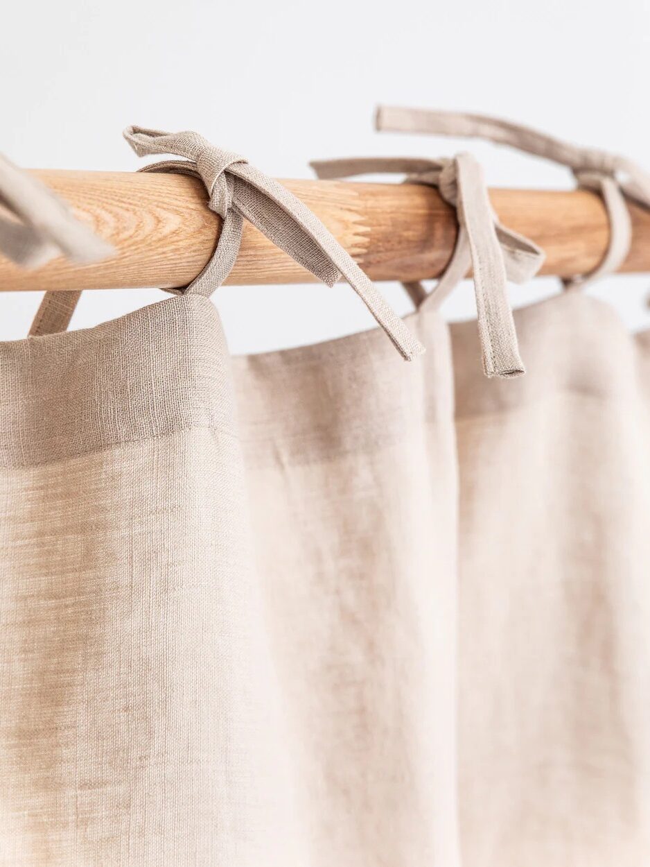 Beige linen curtains tied with fabric ribbons on a wooden rod against a light background.