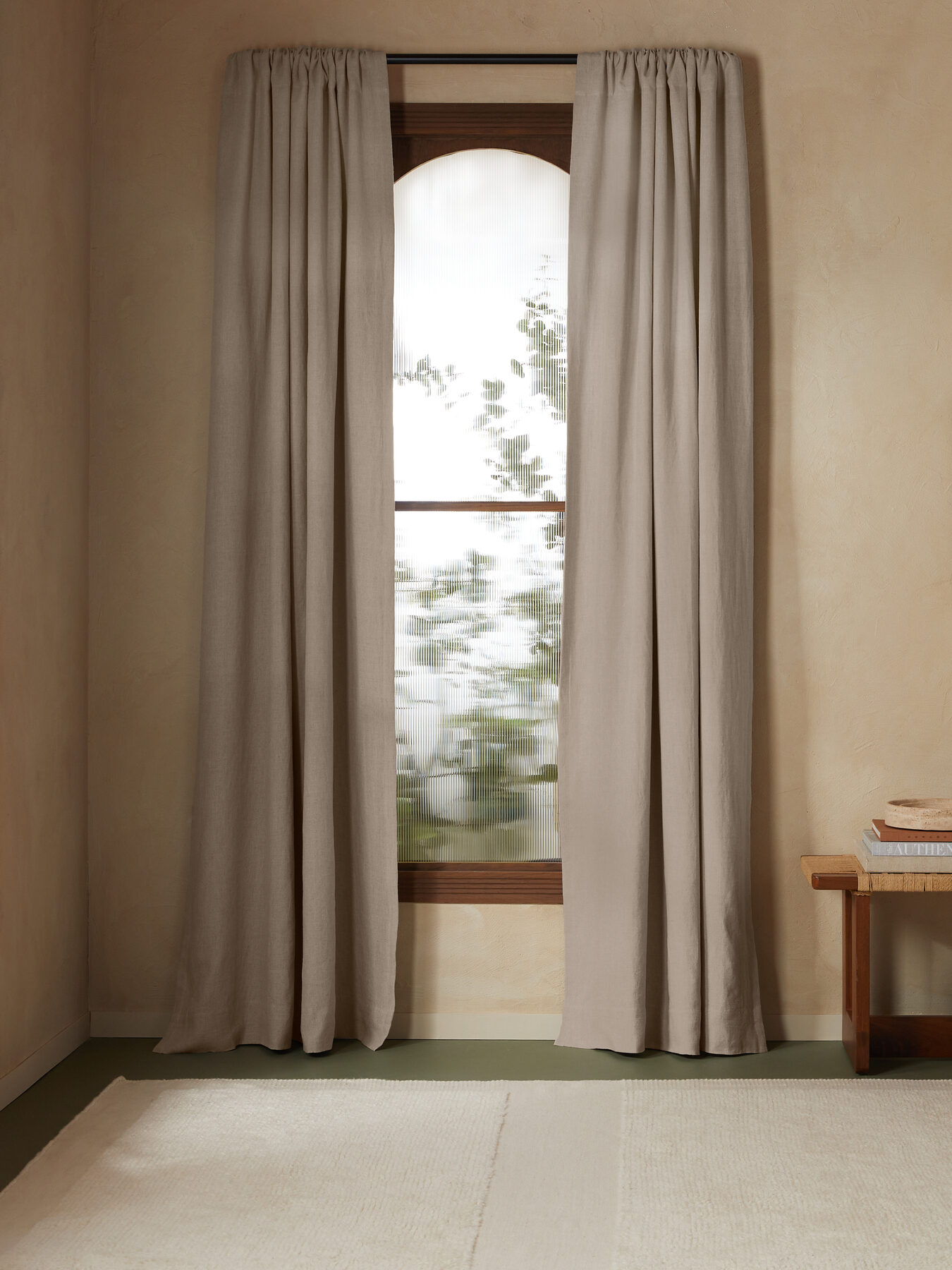 A serene room with beige walls, featuring two curtained windows, a framed landscape painting, and a wooden bench with folded towels.