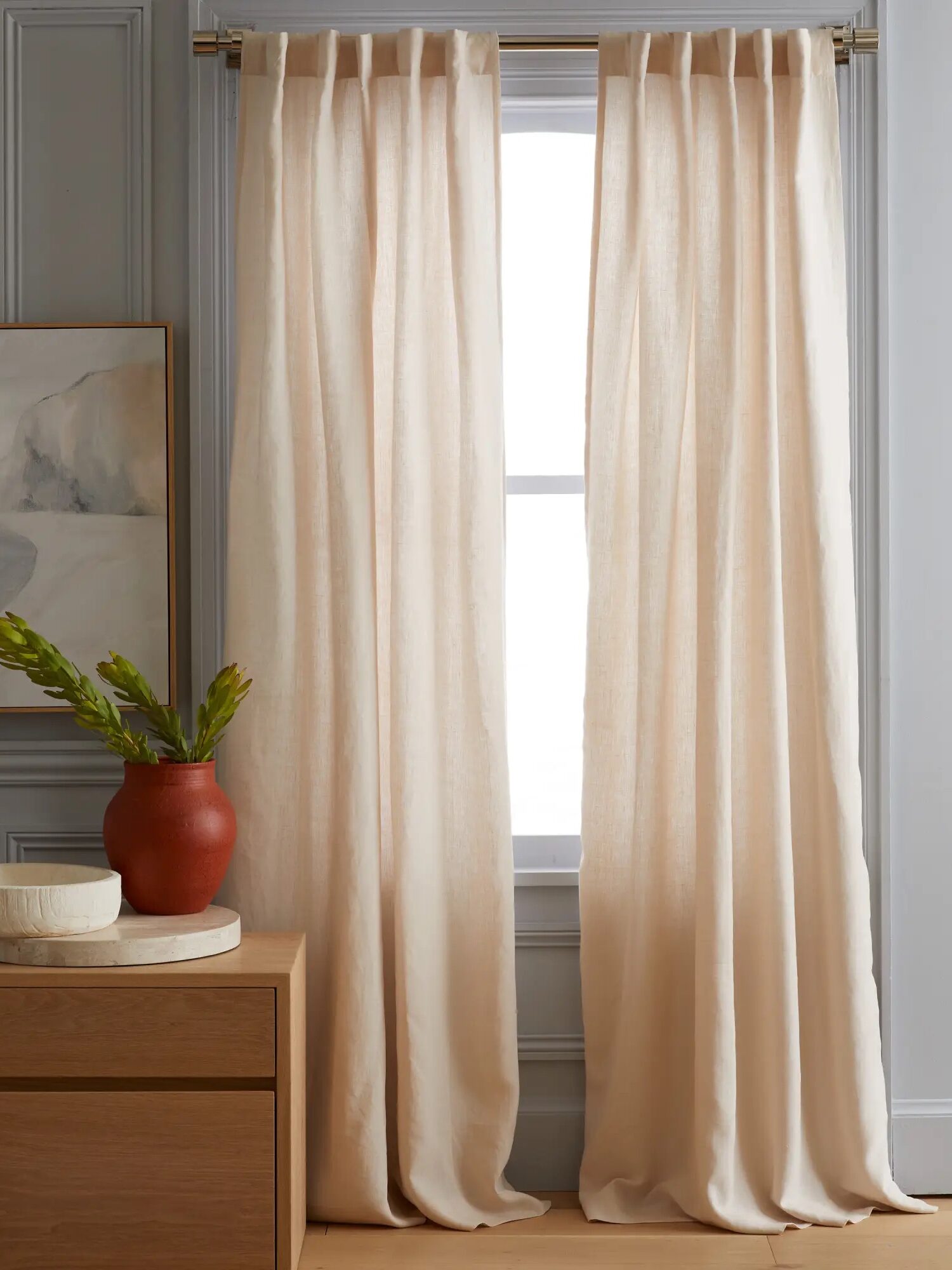 Long beige curtains hang from a rod by large window, with a wooden sideboard and a red vase with green plant on the left.