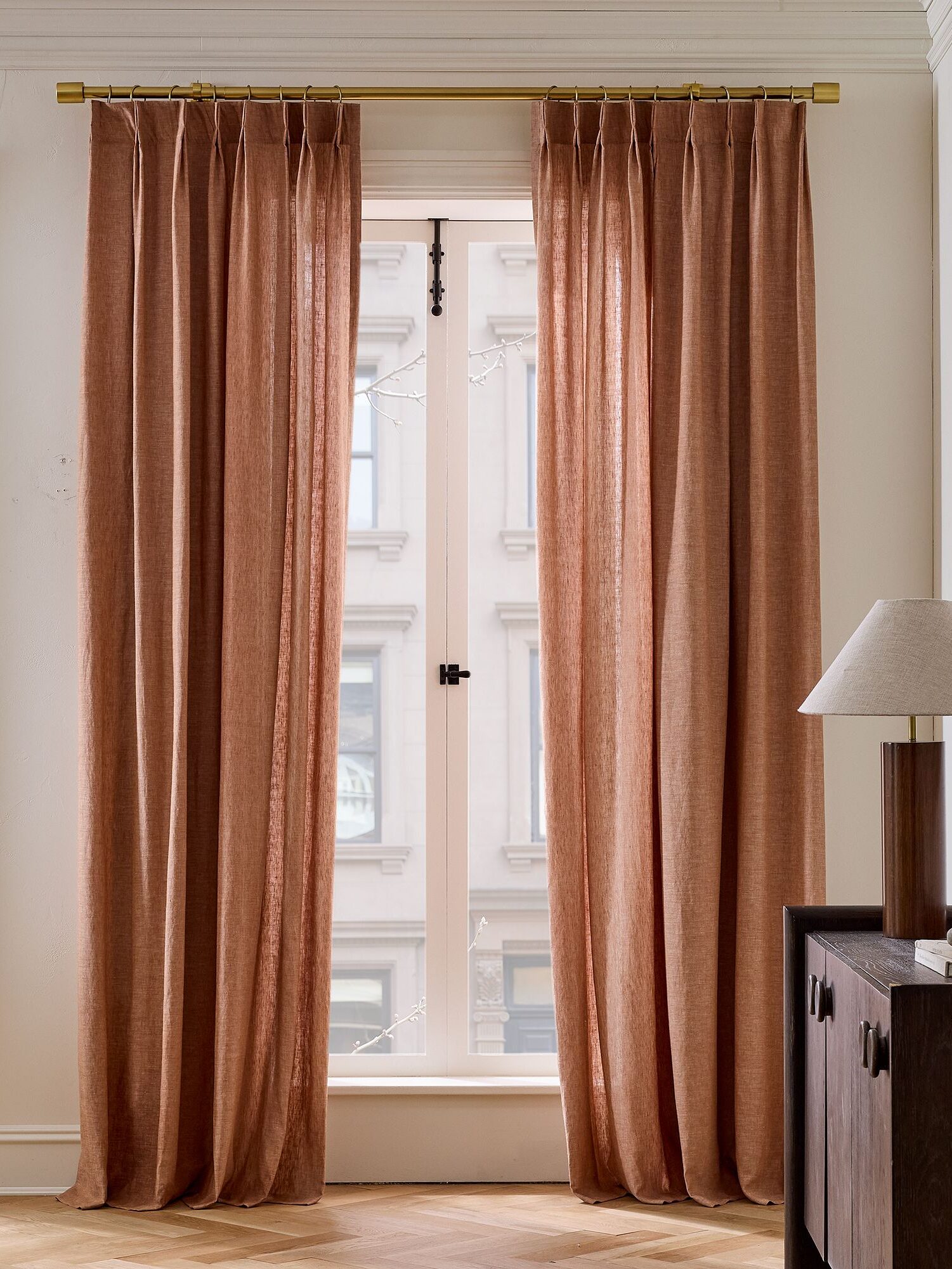 Floor-length terracotta curtains on a gold rod, framing a sunlit window, with a wooden cabinet and lamp on the right.
