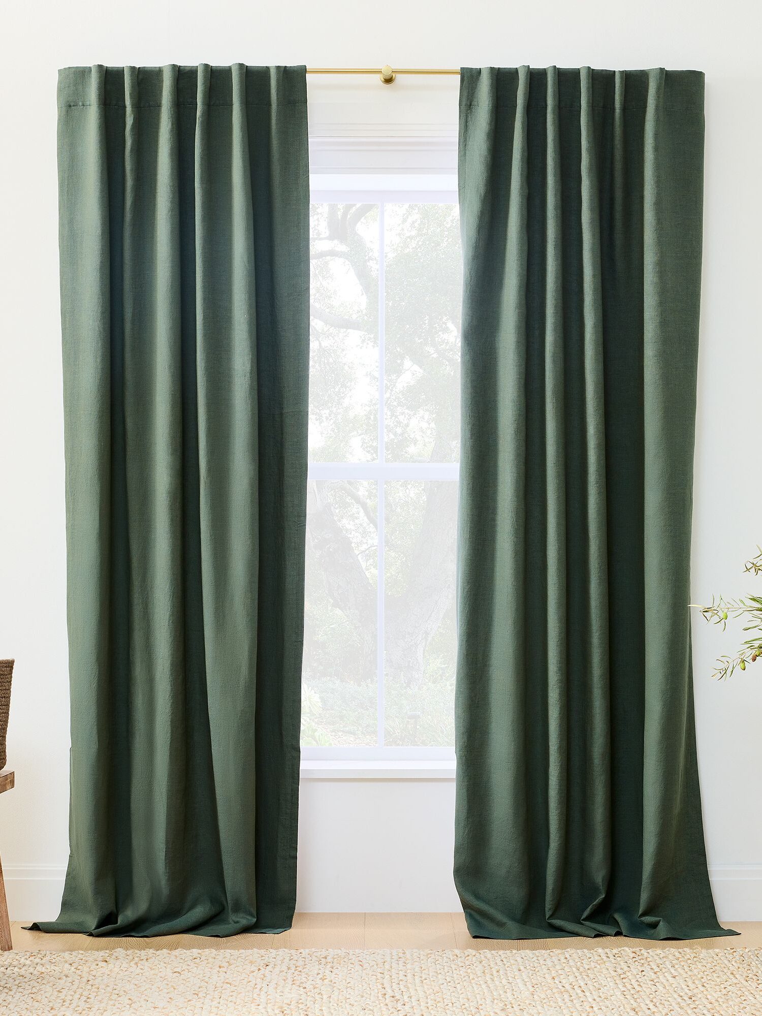 A pair of dark green curtains hangs on a rod, framing a sunlit window in a room with neutral walls and decorative plants.