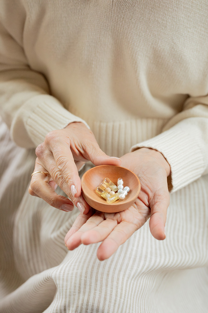 A person in a cream-colored outfit holds a small wooden bowl with assorted supplements in their hands.