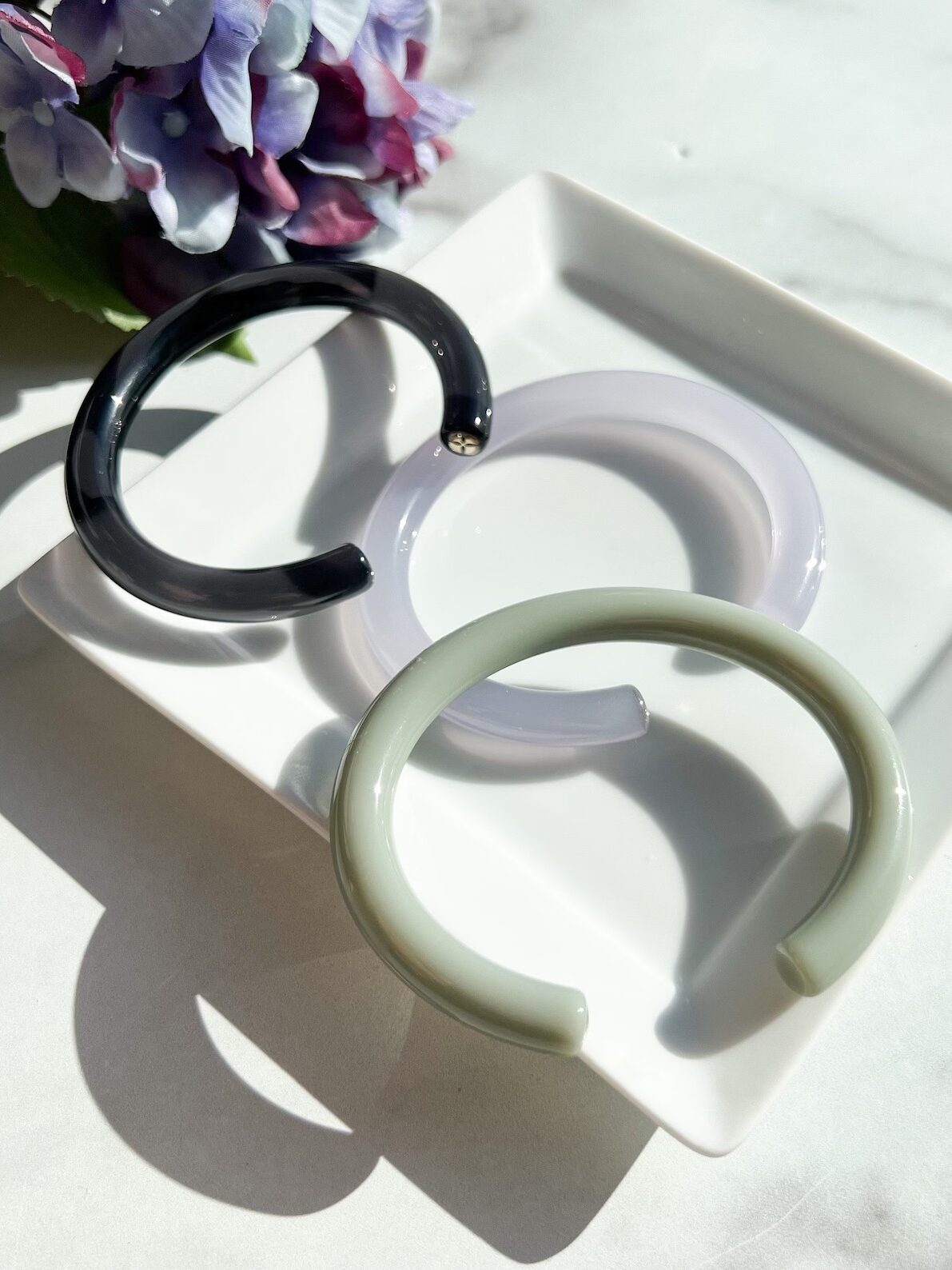 Three acrylic hoop earrings in black, white, and pale green displayed on a square white dish beside a hydrangea flower.