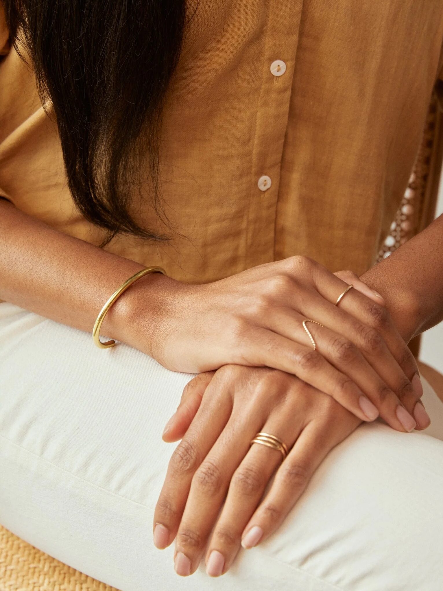 A close-up of a woman's hands resting on her lap, featuring a gold bracelet and rings, with a mustard-colored shirt visible.