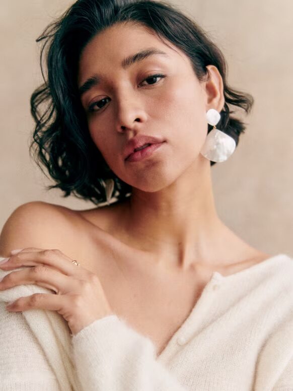 A portrait of a woman with short dark hair, wearing a white sweater and large white earrings, looking thoughtfully at the camera.