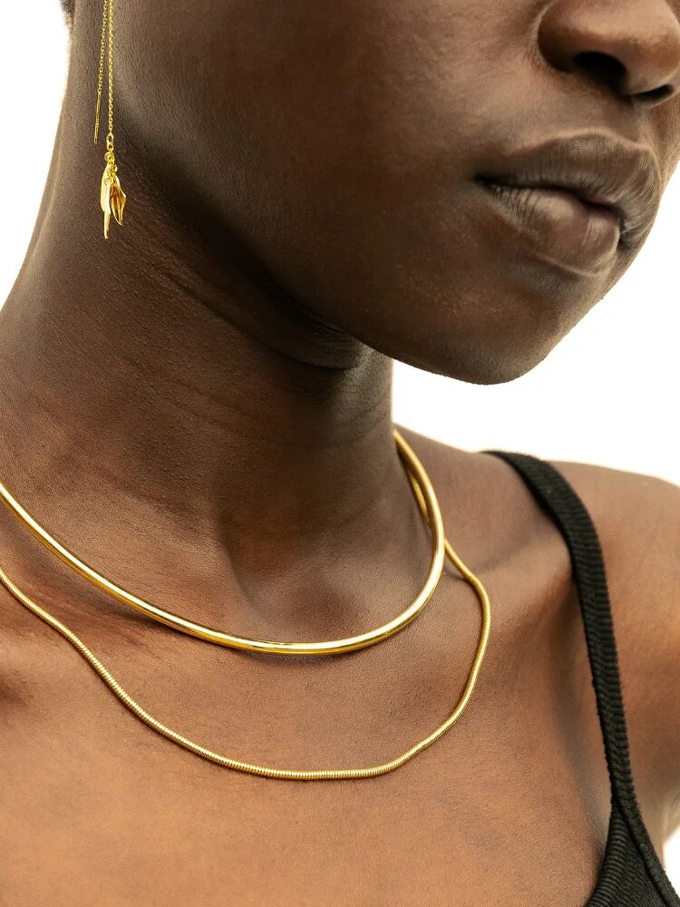 Close-up of a woman wearing gold earrings and necklaces, focusing on her lower face and neck against a white background.