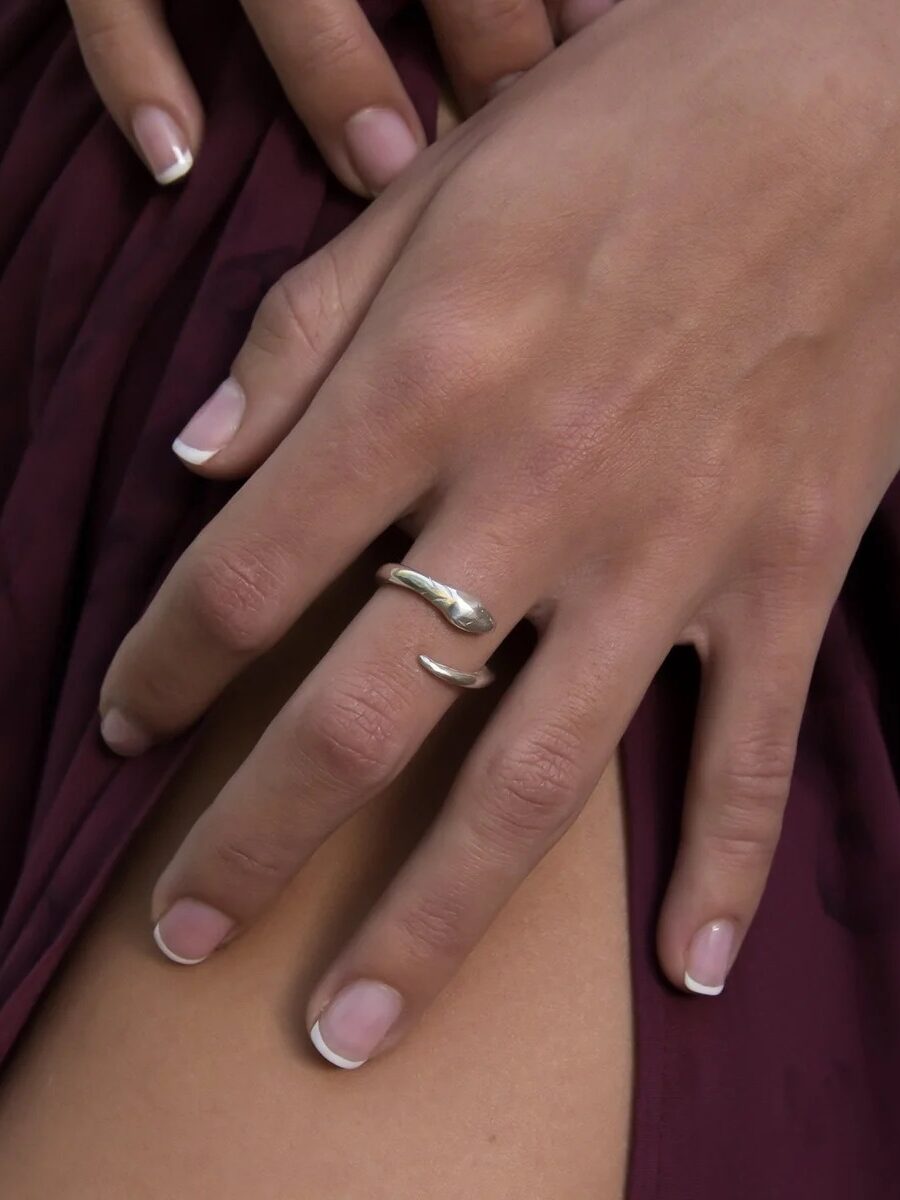 Close-up of a hand with a silver wave ring on the middle finger, resting on another hand against a burgundy fabric background.