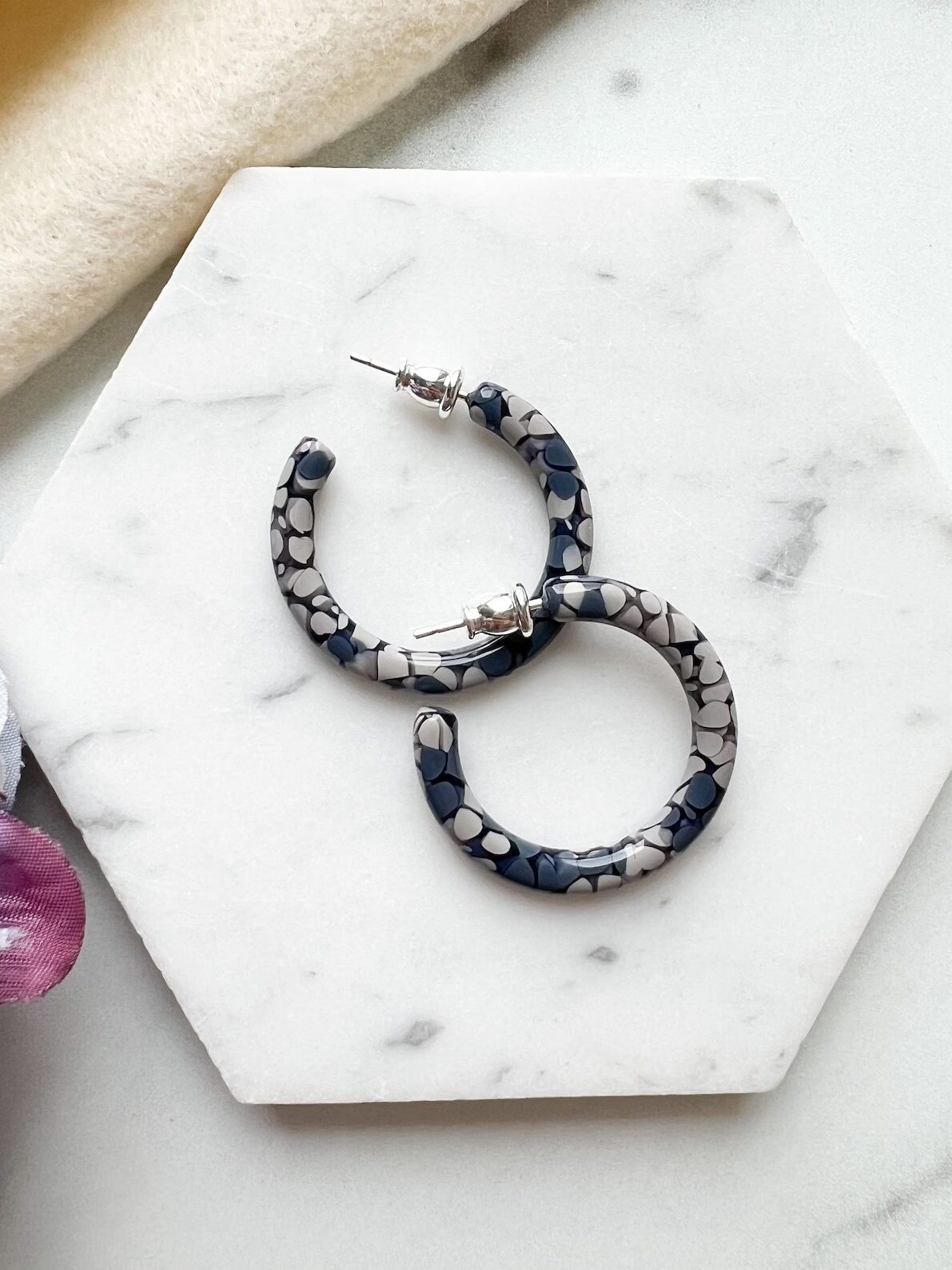 A pair of blue and white patterned hoop earrings rests on a white marble slab next to purple fabric.