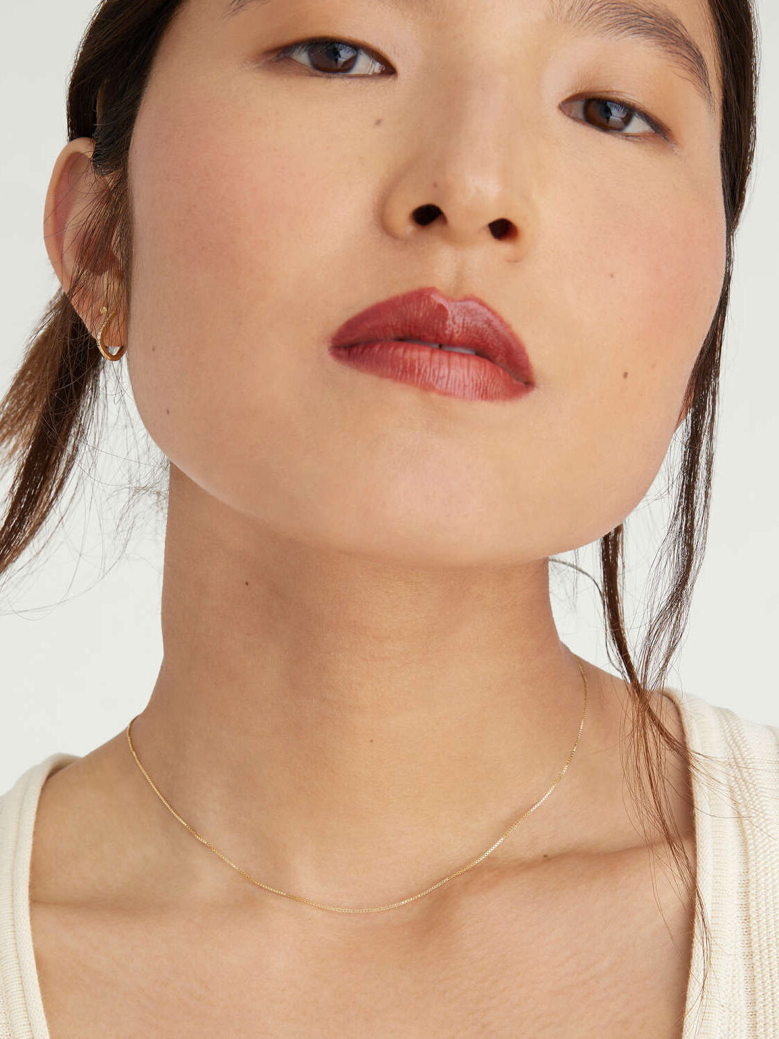 Close-up portrait of a woman with minimal makeup, wearing a delicate gold necklace and a beige top, focusing on her face from the nose up.