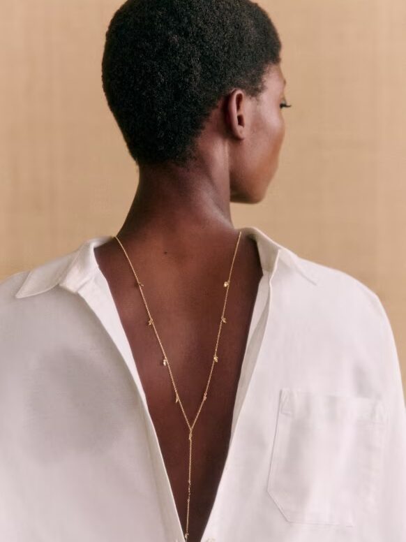 Woman with short hair wearing a white shirt and a delicate gold necklace, viewed from behind against a beige background.