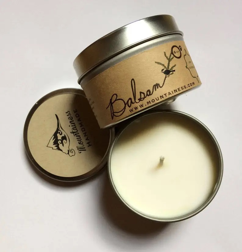A balsam-scented candle in a small tin container with a labeled lid open beside it on a white surface.
