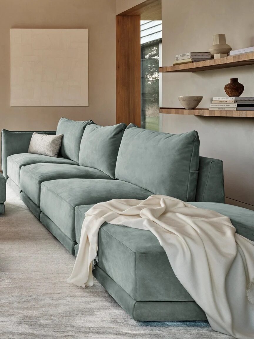 Modern living room with large green sectional sofa, matching armchairs, and a central ottoman. A blanket is draped on the sofa. Shelves with decor items are on the wall; large window lets in natural light.