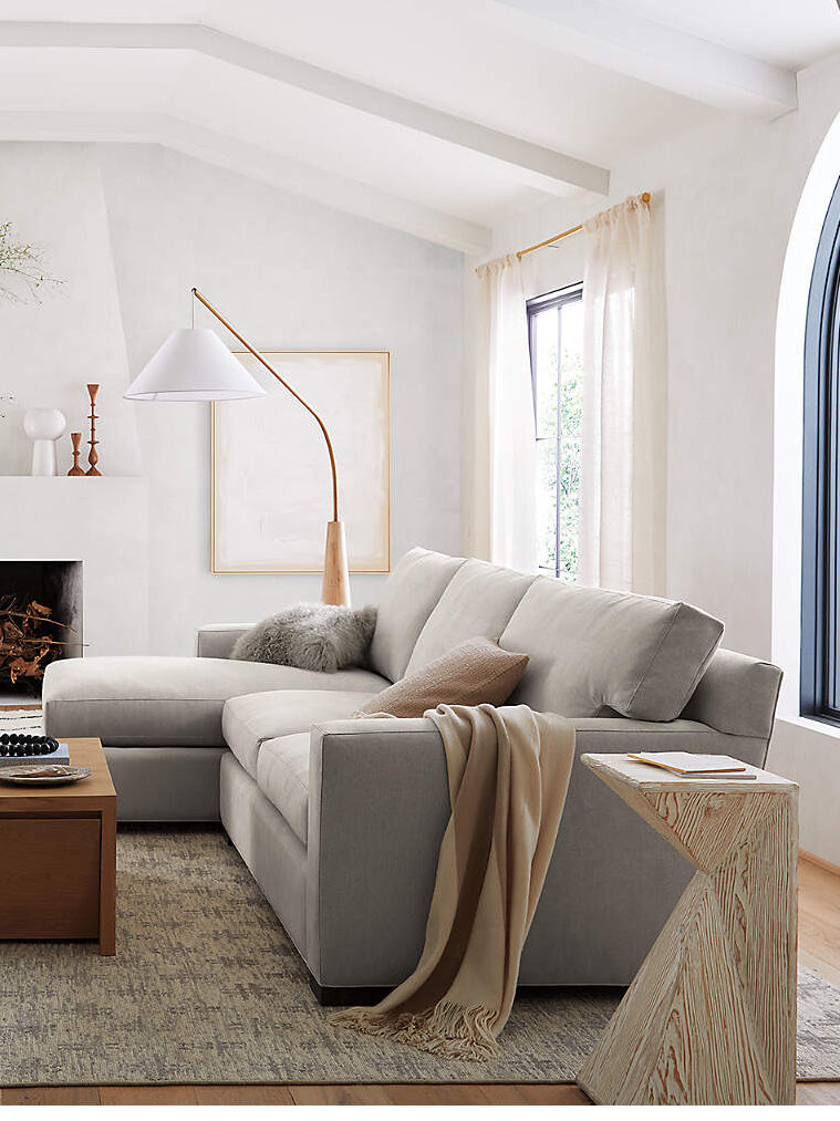 A modern living room with a beige sectional sofa, wooden coffee table, and fireplace. Large windows, light curtains, and a floor lamp complete the minimalist decor. A wicker chair is in the corner.