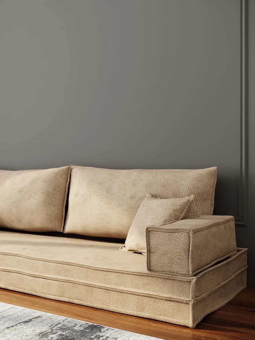 A beige sofa with cushions is placed against a gray wall. A round, wall-mounted light is on the left side above the sofa. A white and gray rug covers the wooden floor partially.
