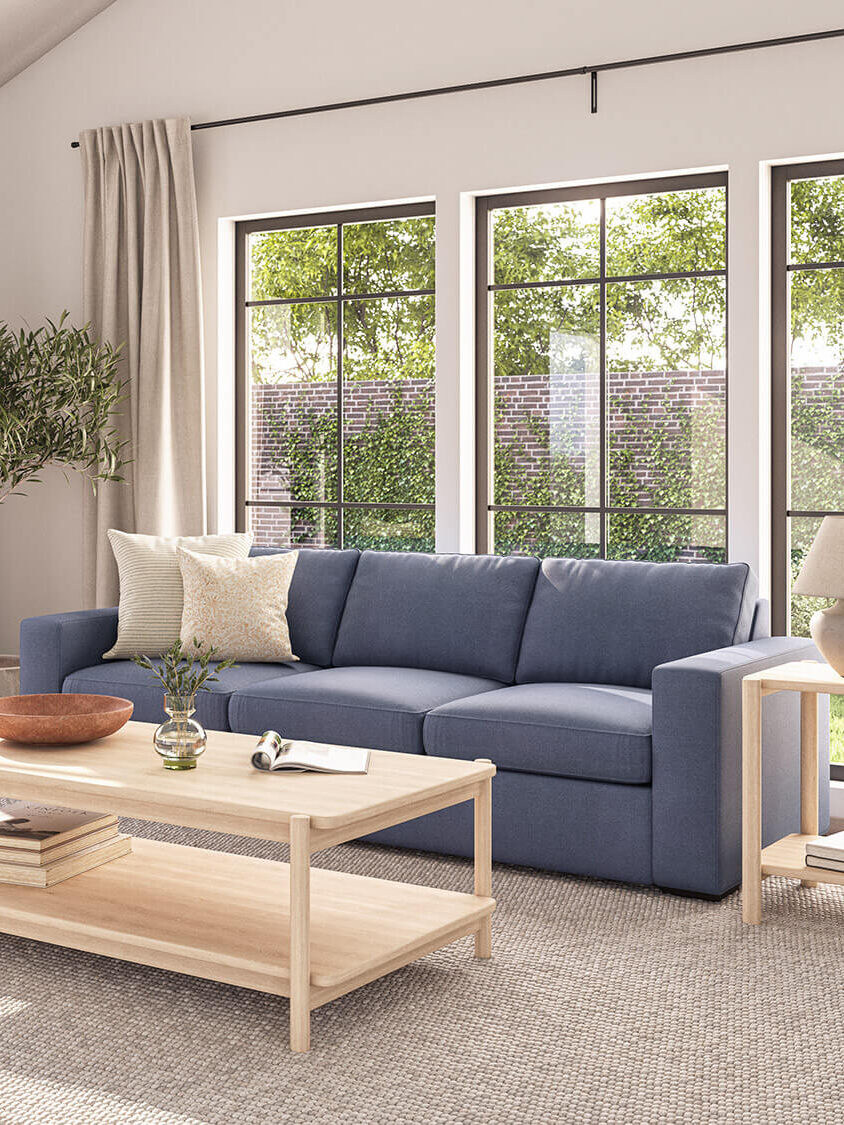 A bright living room with a blue sofa, wooden coffee table, armchair, lamp, plant, and large windows showing an outdoor garden. Neutral tones and natural light create a cozy atmosphere.
