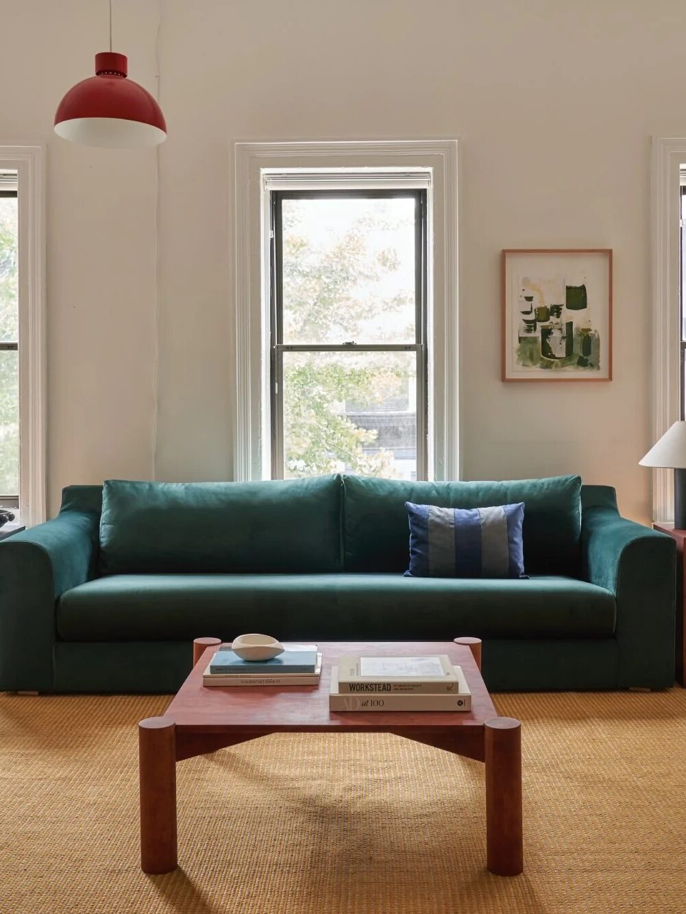 A living room with a green sofa, a wooden coffee table, a white armchair, and three windows. The room features a red pendant light, a beige rug, and a framed plant artwork on the wall.