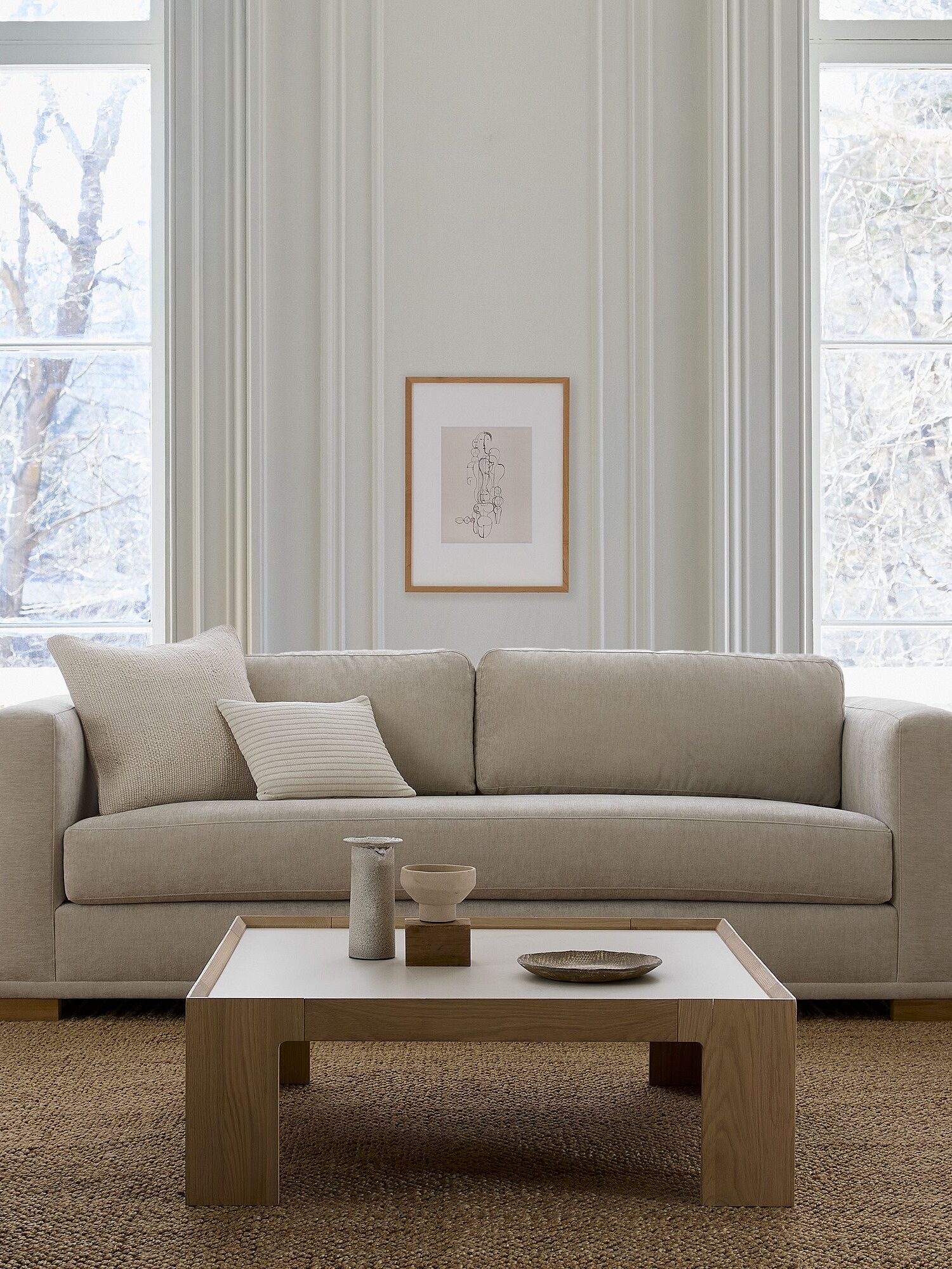 A minimalist living room features a beige sofa with white pillows, a wooden coffee table with decor, and large windows showcasing a snowy outdoor view.