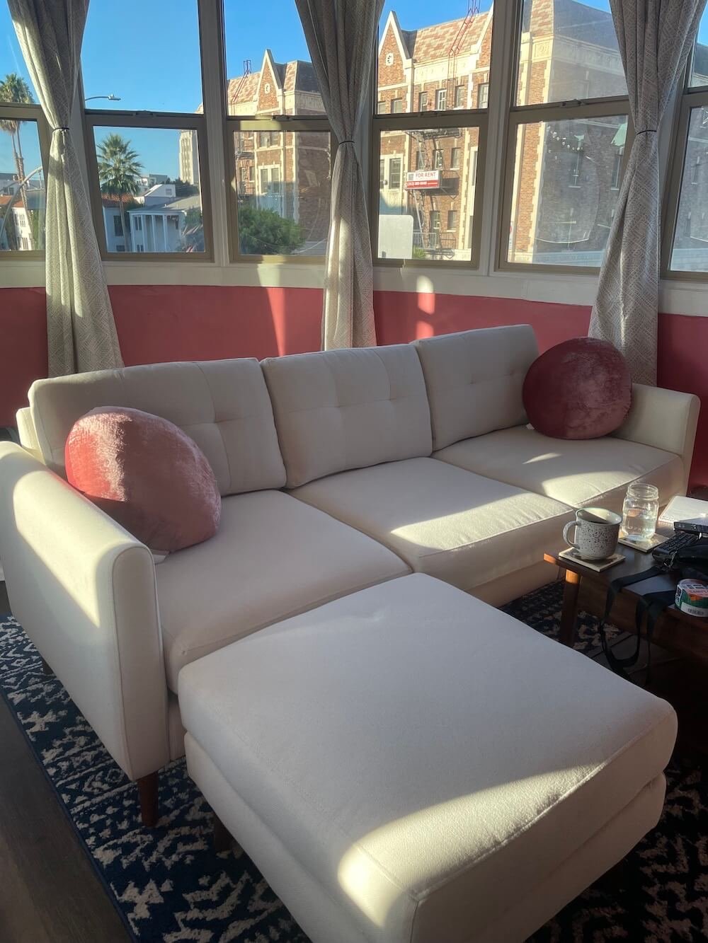 A beige sectional sofa with two pink round pillows from Burrow is placed in a TGT editor's sunlit room with red-painted walls and large windows showing a view of buildings outside. A small table with items sits next to the sofa.