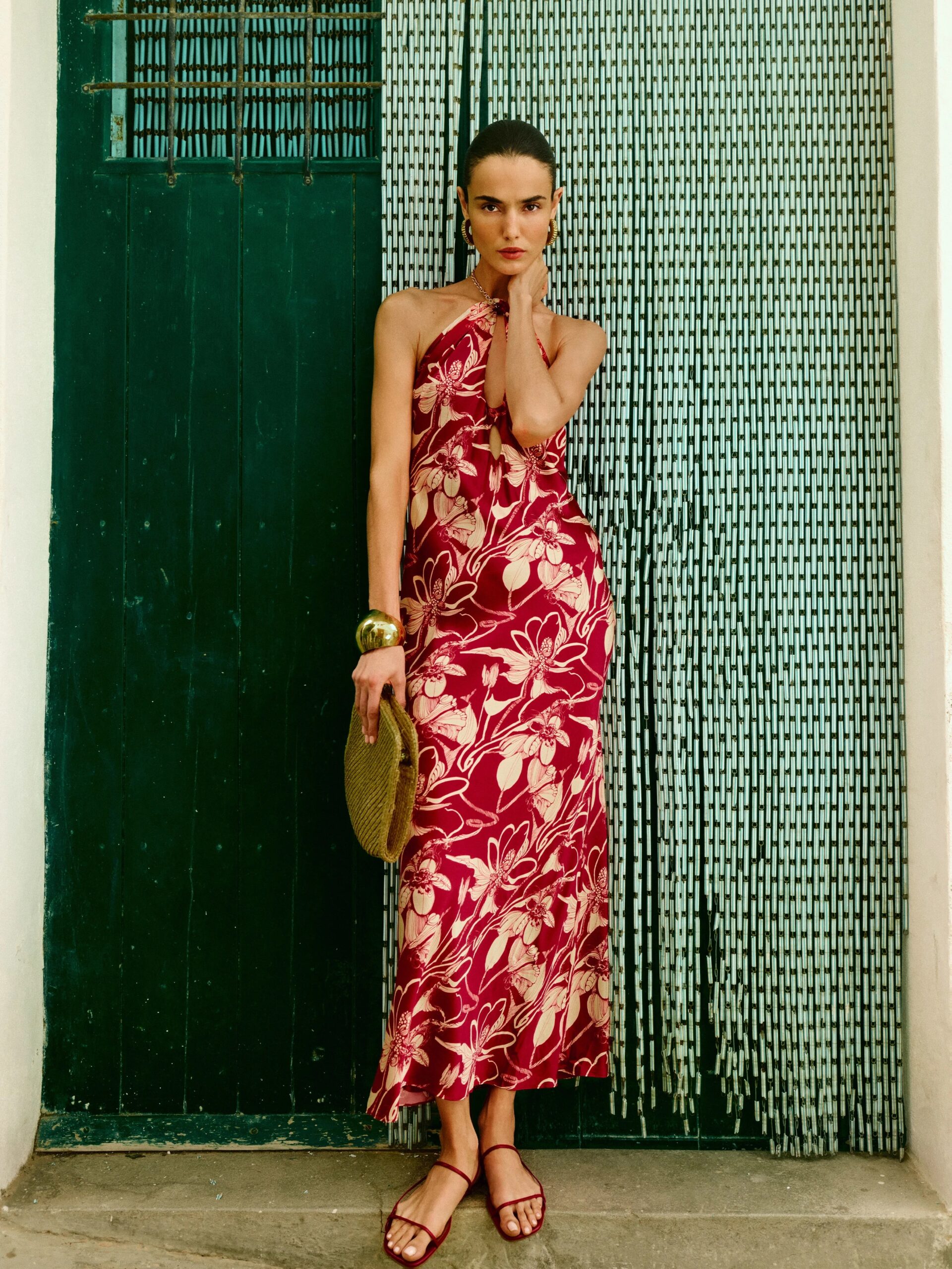 A woman in a patterned, sleeveless dress stands against a green door, holding a woven handbag and wearing bracelets.