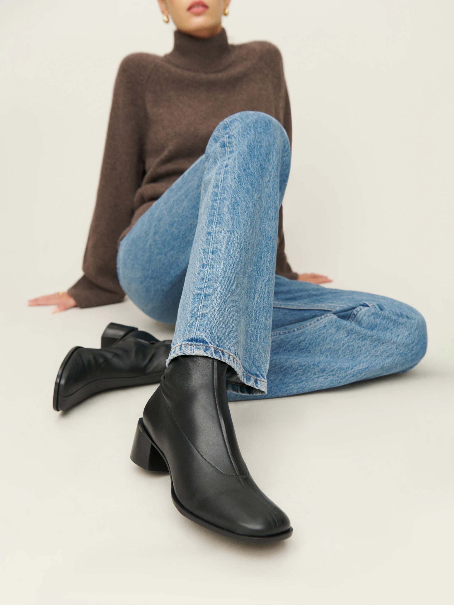 Person wearing a brown sweater and blue jeans, sitting on the floor, showcasing black ankle boots with block heels.