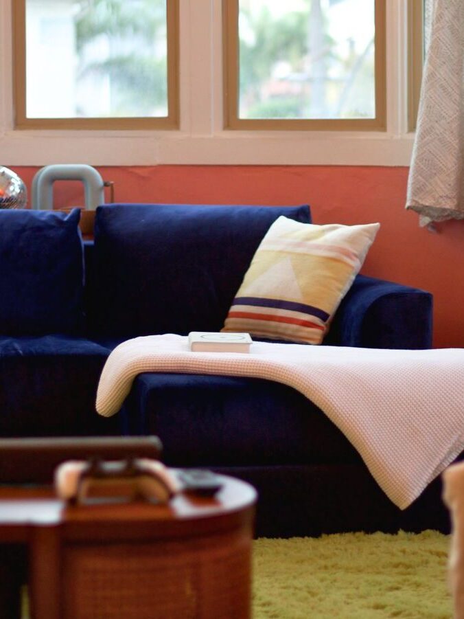 A cozy living room with a blue couch, a white blanket, and a patterned pillow. A book is on the couch, a bike is parked nearby, and large windows provide natural light.