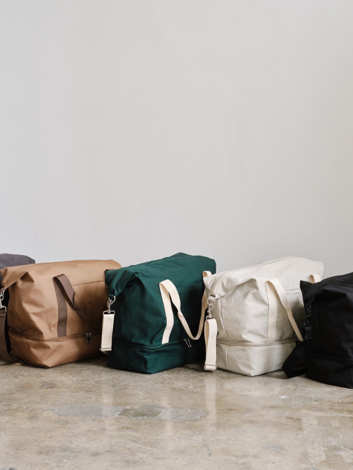 Five duffel bags in various colors (gray, tan, green, white, black) aligned on a concrete floor against a neutral wall.