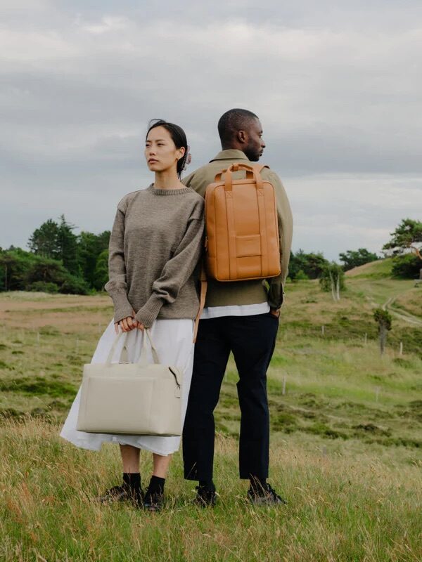 A woman and a man standing back-to-back in a grassy field, the woman holding a white bag and the man wearing a tan backpack.