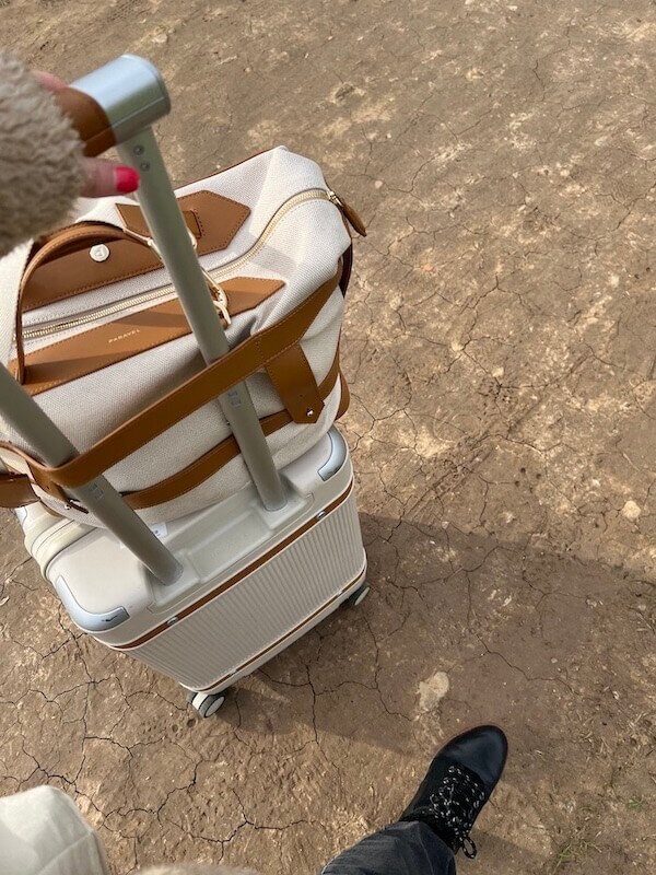 A person stands on a dirt path next to a white and tan suitcase with an extended handle, showing one black shoe in the frame.