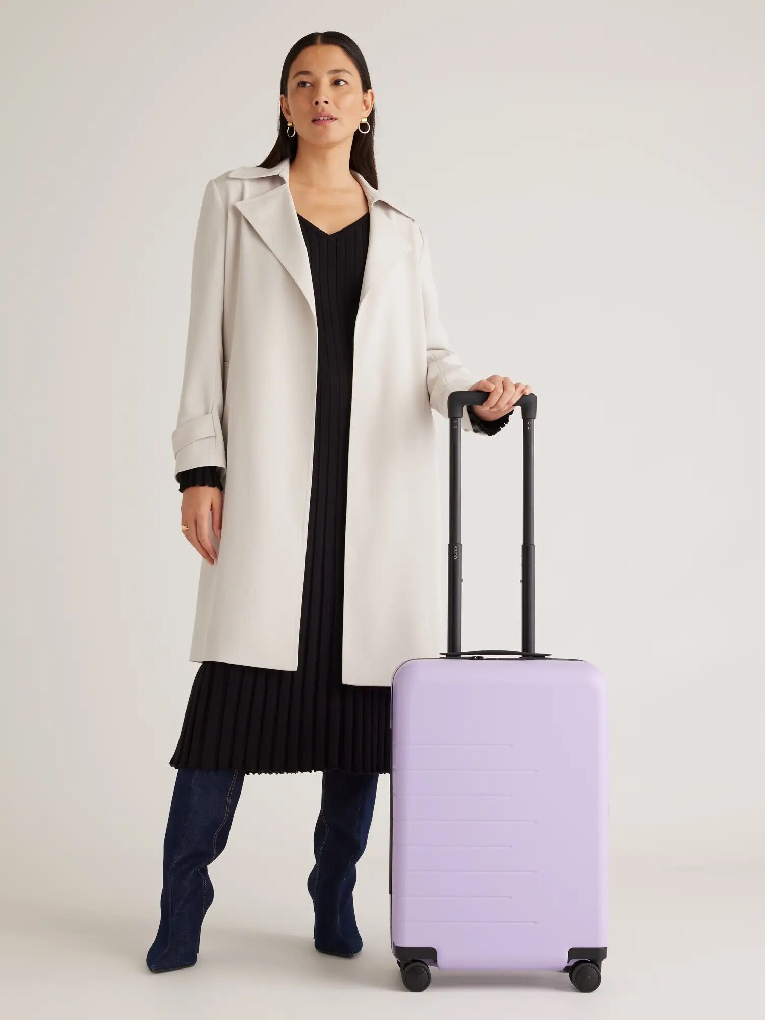 Woman in a stylish coat and boots standing with a purple suitcase, against a plain background.