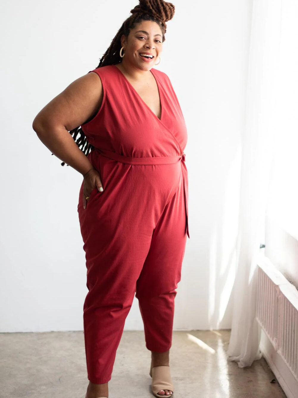 A smiling woman in a red jumpsuit posing confidently with one hand on her hip in a bright, white room.
