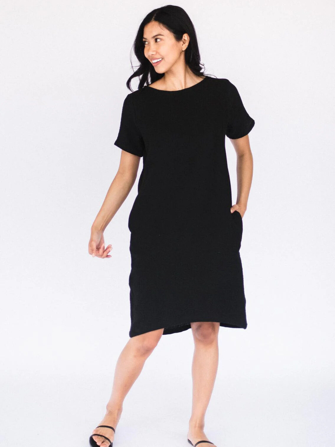A woman in a black dress and sandals smiling and looking to the side against a white background.