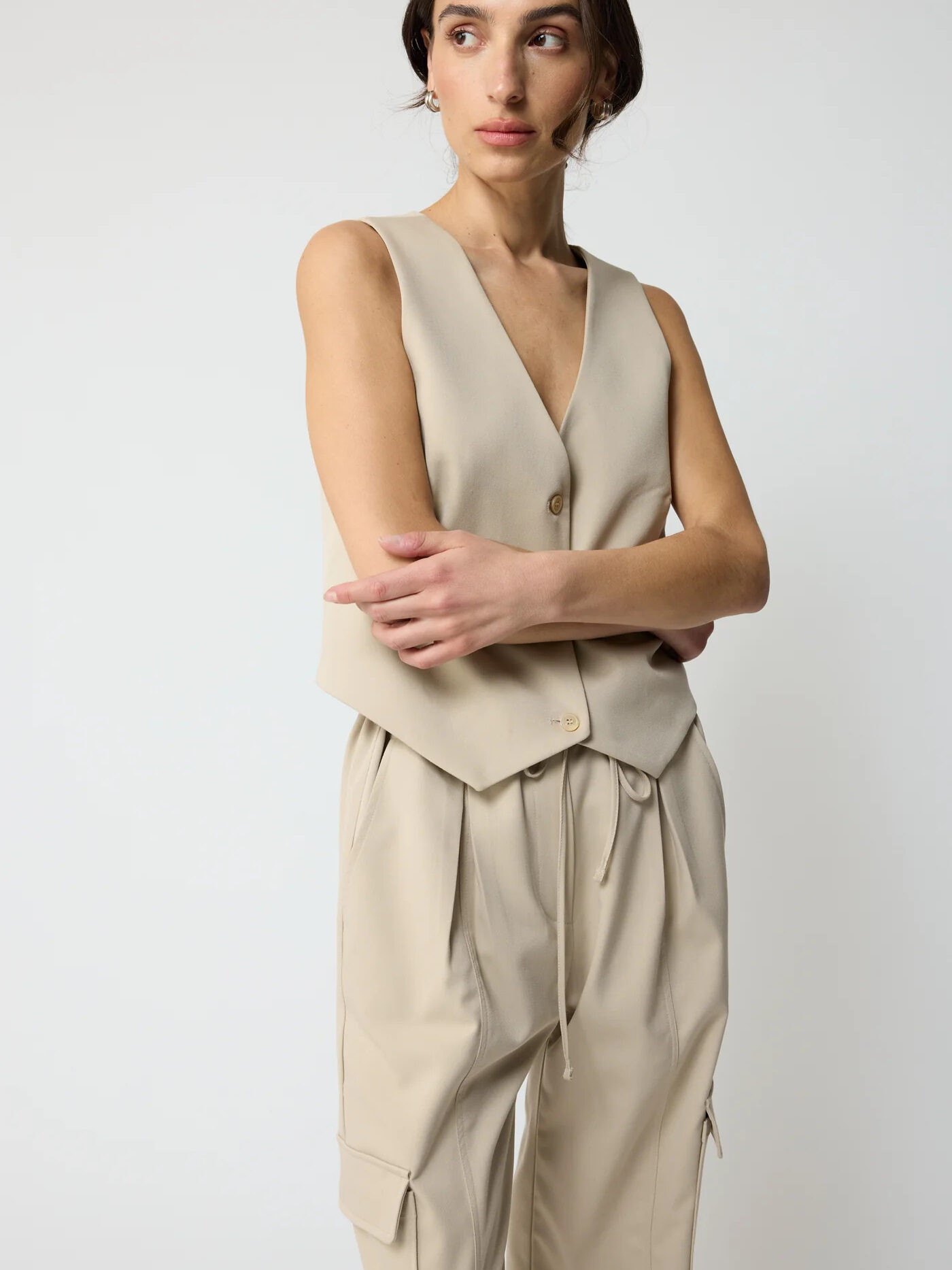 Woman in a beige sleeveless top and matching trousers posing with crossed arms on a plain background.