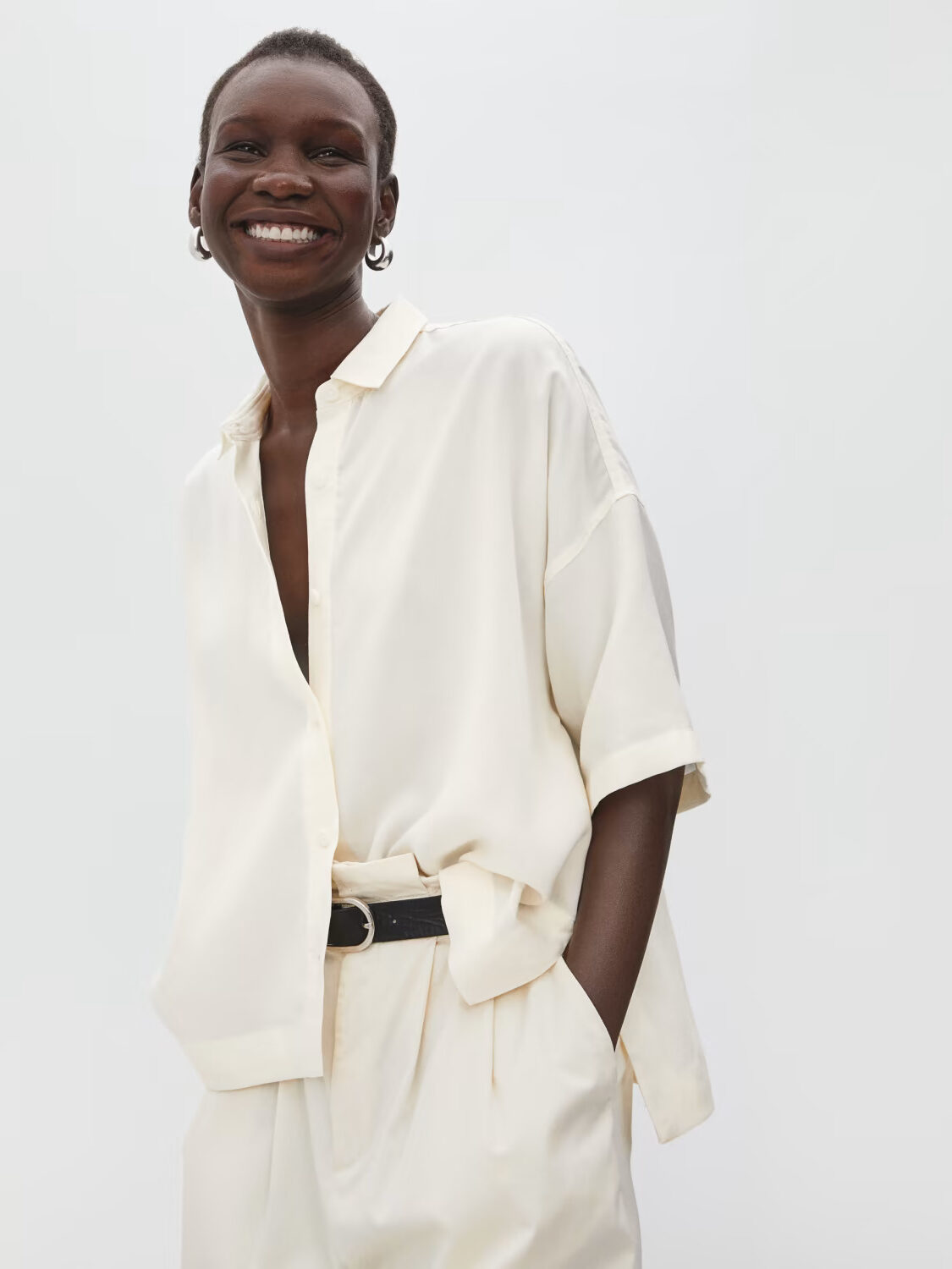 Fashionable woman in an ivory shirt and trousers, smiling at the camera with a minimalist white background.