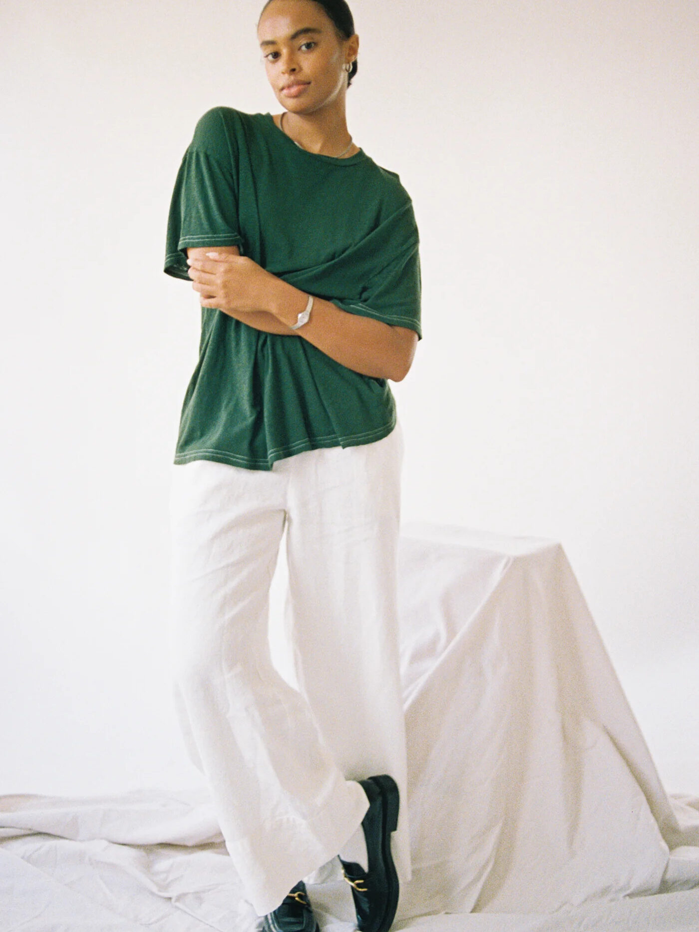 A person in a green t-shirt and white pants stands confidently against a plain backdrop, with one arm crossed.