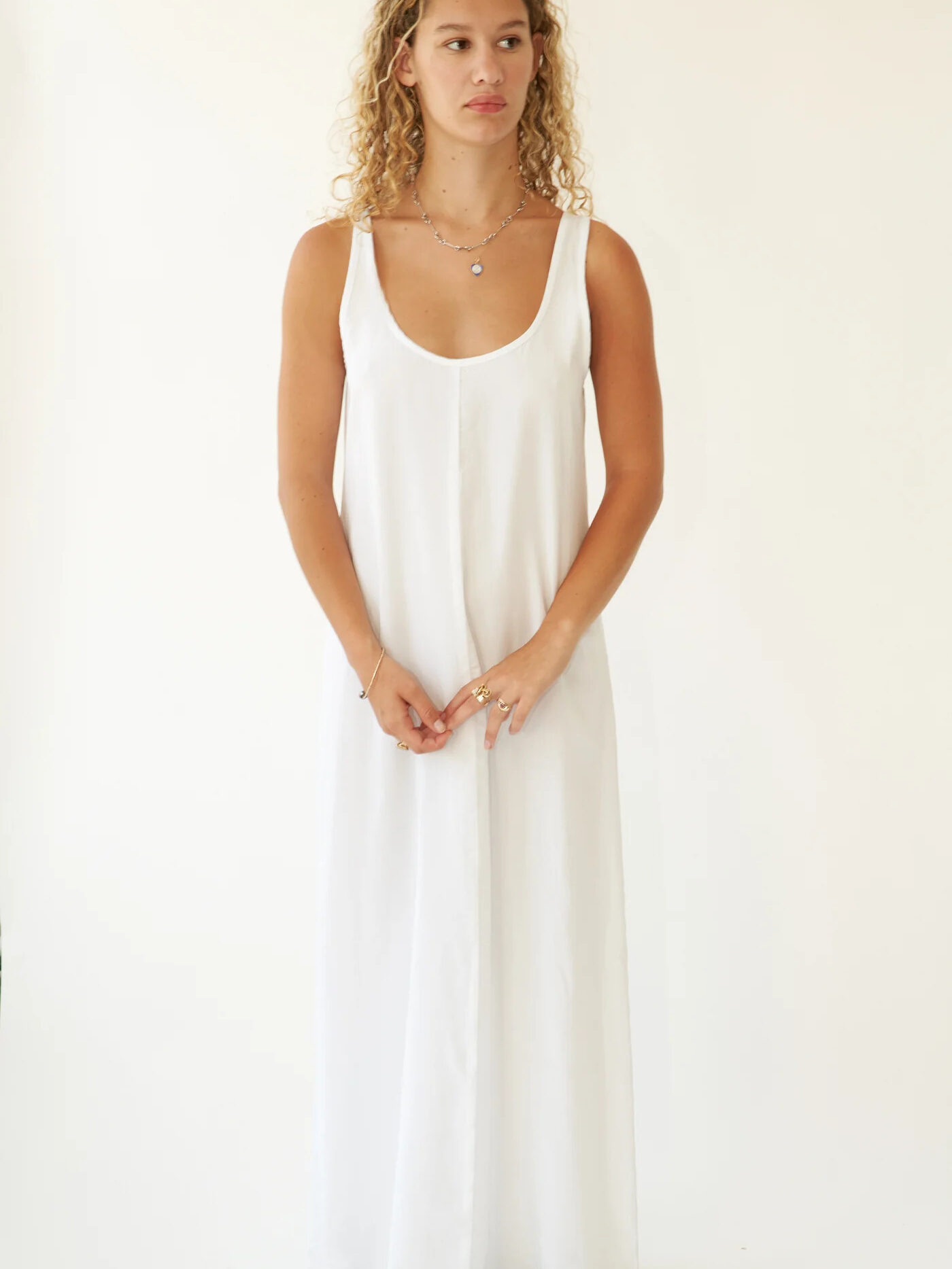 A young woman with curly hair, wearing a simple long white dress, stands against a white background, looking slightly to the side.